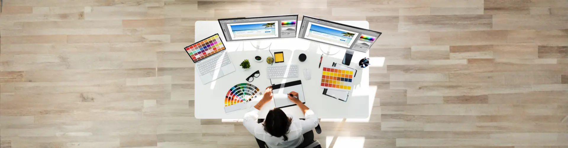 Das Bild zeigt einen Arbeitsplatz mit mehreren Monitoren, Farbmusterkarten und Grafiktablett – es steht für kreative Arbeit in Design oder Mediengestaltung.