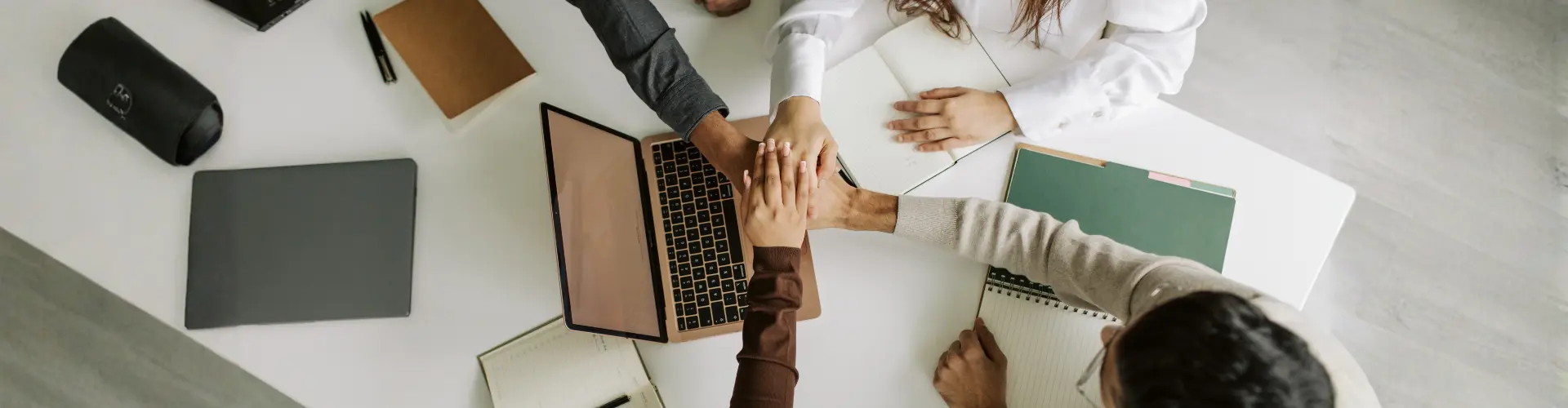 Das Bild zeigt mehrere Personen, die am Tisch ihre Hände übereinanderlegen – es steht für Teamwork, Zusammenhalt und gemeinsames Arbeiten.