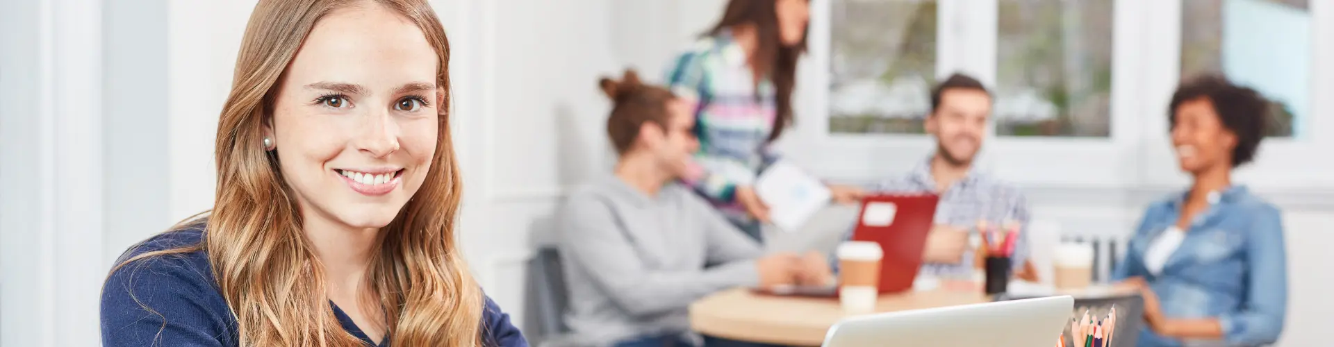 Junge Frau sitzt vor einen Laptop im Hintergrund sind weitere Personen an einem Tisch