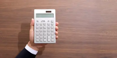 Das Bild zeigt eine Hand mit Taschenrechner vor Holz-Hintergrund – es steht für Steuern und Abgaben in der Ausbildung.