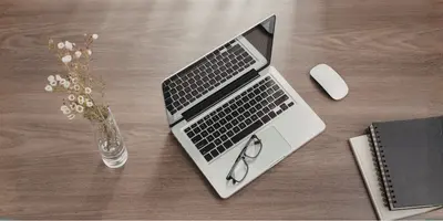 Das Bild zeigt einen aufgeräumten Schreibtisch mit Laptop, Notizbüchern, Maus und einer Vase mit Blumen – es steht für einen organisierten Arbeitsplatz und konzentriertes Arbeiten.