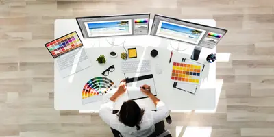 Das Bild zeigt einen Arbeitsplatz mit mehreren Monitoren, Farbmusterkarten und Grafiktablett – es steht für kreative Arbeit in Design oder Mediengestaltung.