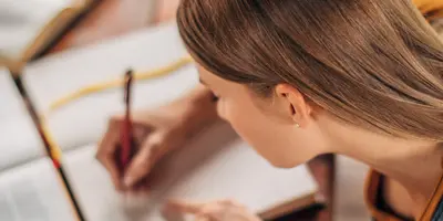 Das Bild zeigt eine Person, die konzentriert am Schreibtisch sitzt und in Bücher schreibt – es steht für Lernen.