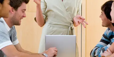 6 Personen sitzen an einen Tisch und eine Frau steht vorne und erklärt etwas