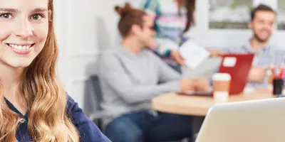 Junge Frau sitzt vor einen Laptop im Hintergrund sind weitere Personen an einem Tisch