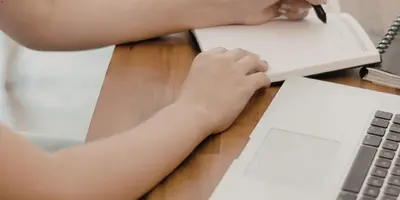 Person sitzt am Schreibtisch und schreibt etwas auf daneben steht ein Laptop